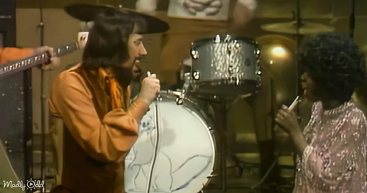A man in a hat and long coat stands in front of a drum set, holding a microphone, with another person in the background.