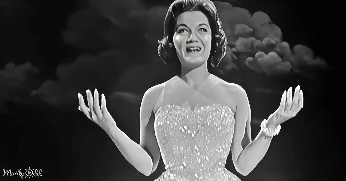 A woman in a sparkling dress with her hands outstretched, smiling, against a dark, cloud-filled background.
