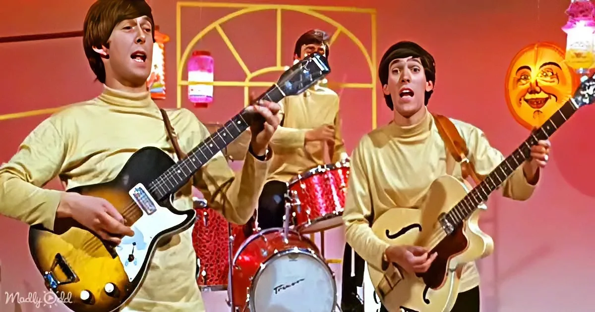Musicians in beige outfits play guitars on stage with a red drum set and whimsical background.
