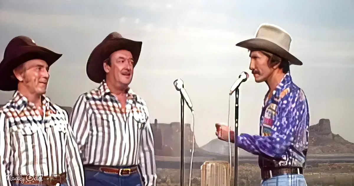 A man in a colorful shirt speaks into a microphone, flanked by two others in cowboy hats, against a desert backdrop.