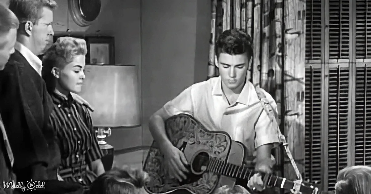 A man plays guitar while others watch, in a vintage black-and-white setting.