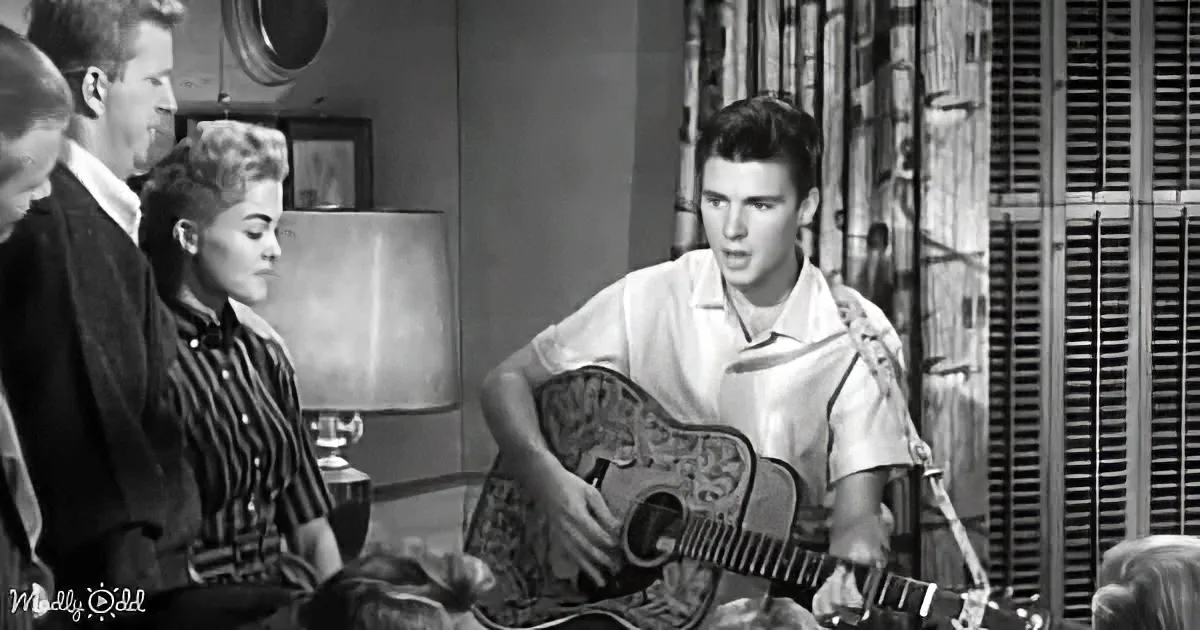 A young man plays guitar in a room with others watching, a lamp and window shutters in the background.