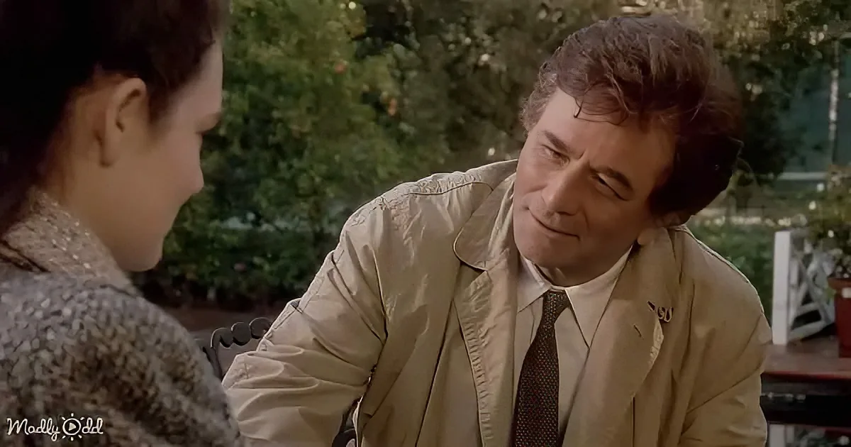 A man in a beige jacket and tie leans forward, looking down at a woman in a patterned coat, both seated outdoors.