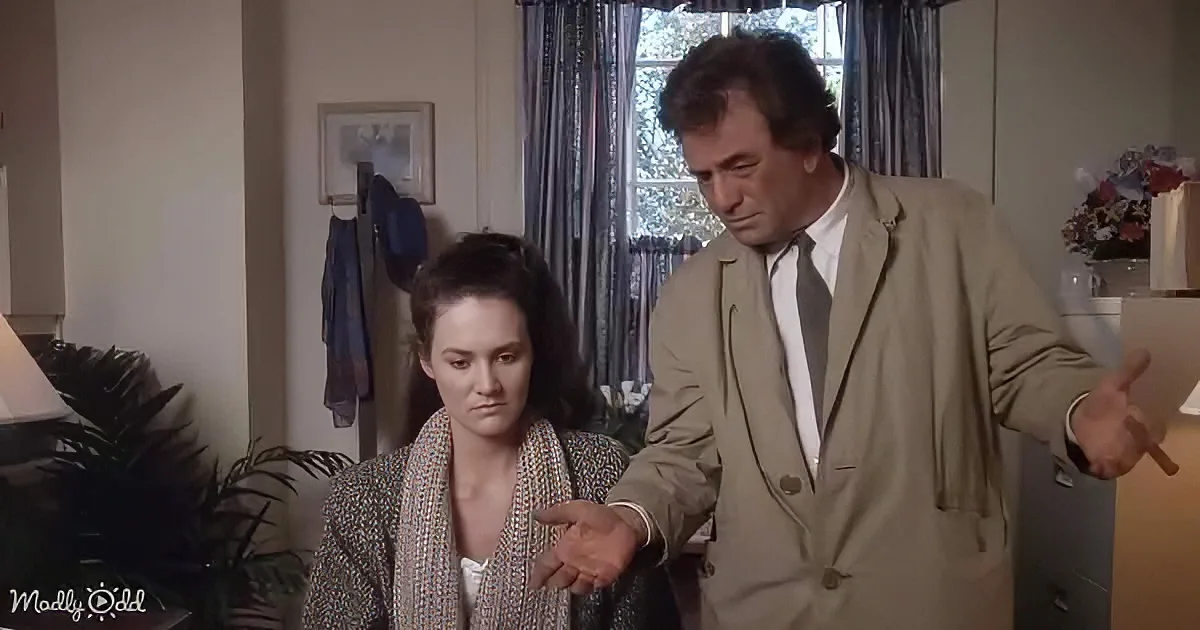 A man in a beige suit gestures emphatically while a woman in a robe looks on, both in a room with plants and curtains.