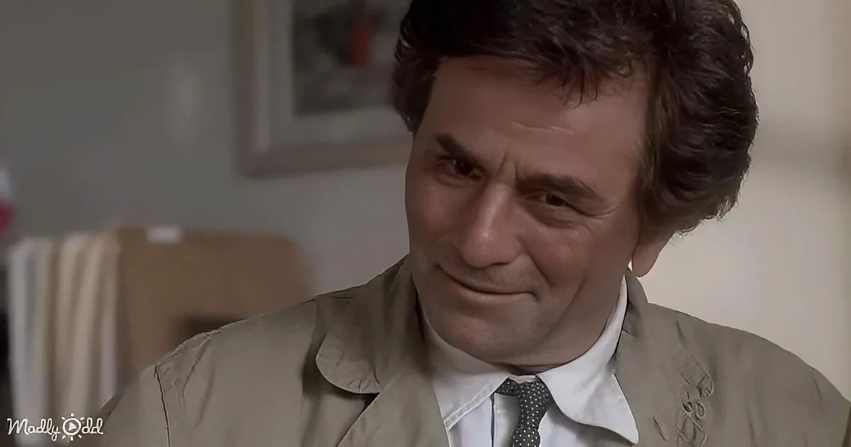 A man in a beige suit and tie is smiling, looking downward, with a framed picture in the background.