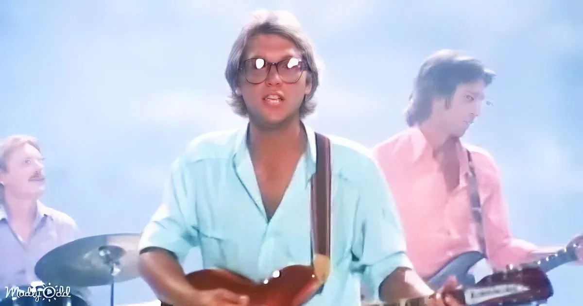 A man in a light blue shirt plays a guitar, wearing glasses, with a band in the background under a blue sky.