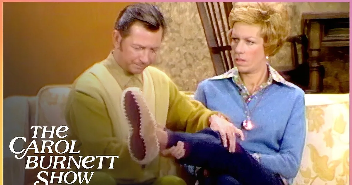 A man in a yellow suit shakes hands with a woman in a blue floral shirt in a warmly lit room.
