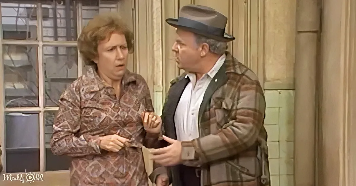 A man in a hat and plaid jacket holds a woman's arm, both appearing surprised or concerned.