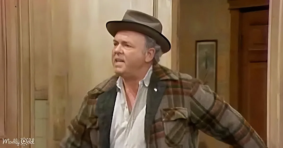 A man in a plaid jacket and fedora stands indoors, looking upwards with a surprised expression.