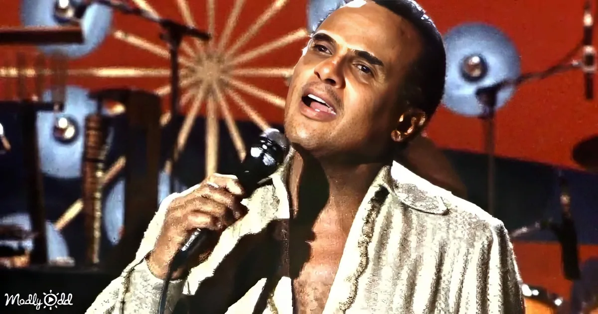 A man in a white shirt sings into a microphone on stage with a drum set in the background.