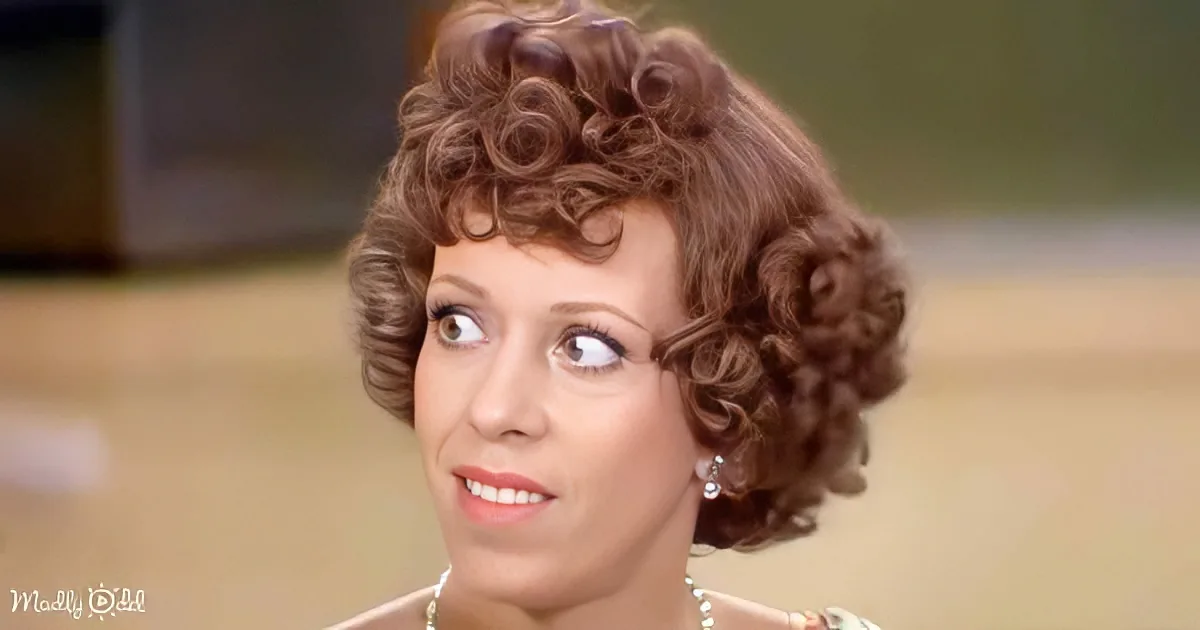 A woman with curly brown hair, wearing pearl earrings and a necklace, smiles while looking to the side.