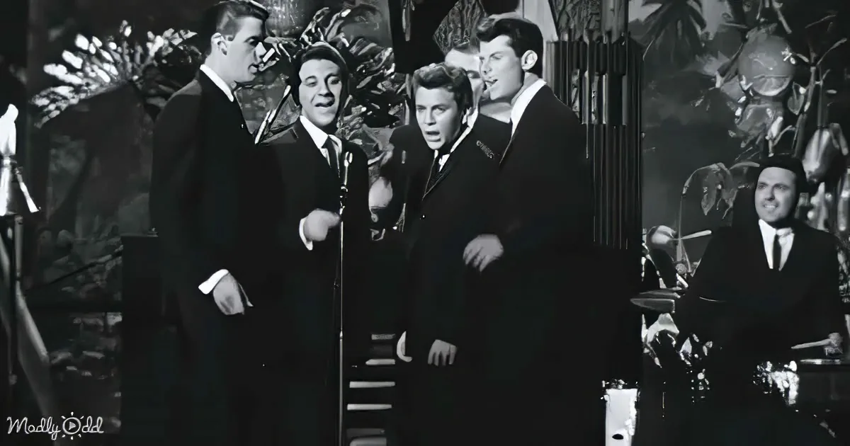 Four men in suits sing on stage, one playing drums in the background.