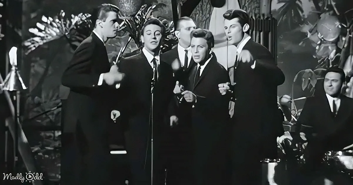 A group of men in suits sing on stage with microphones, surrounded by a band and tropical decorations.