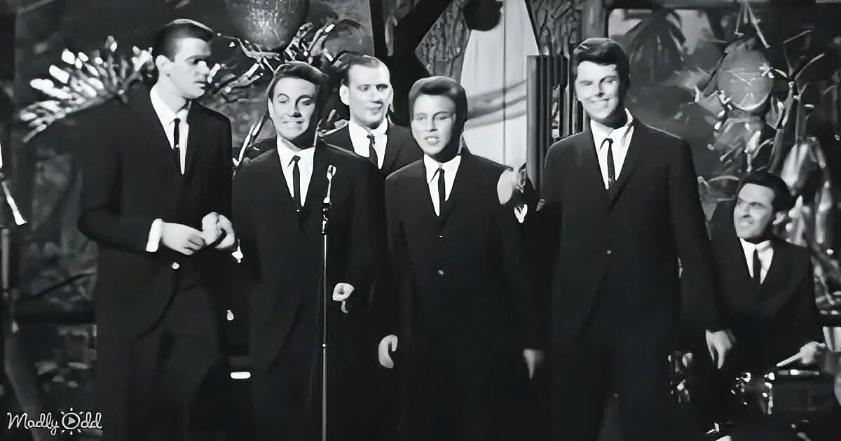 A group of men in suits stands on stage, singing into microphones, with a drummer in the background.