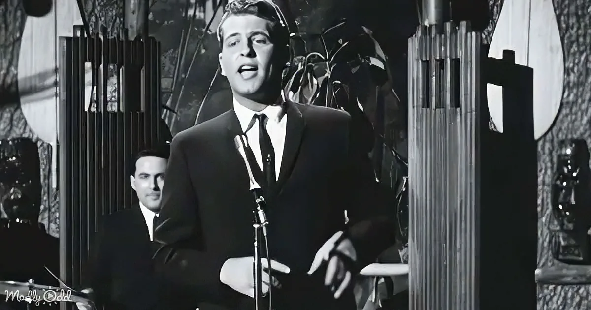 A man in a suit sings into a microphone, surrounded by musical instruments and a drummer in the background.