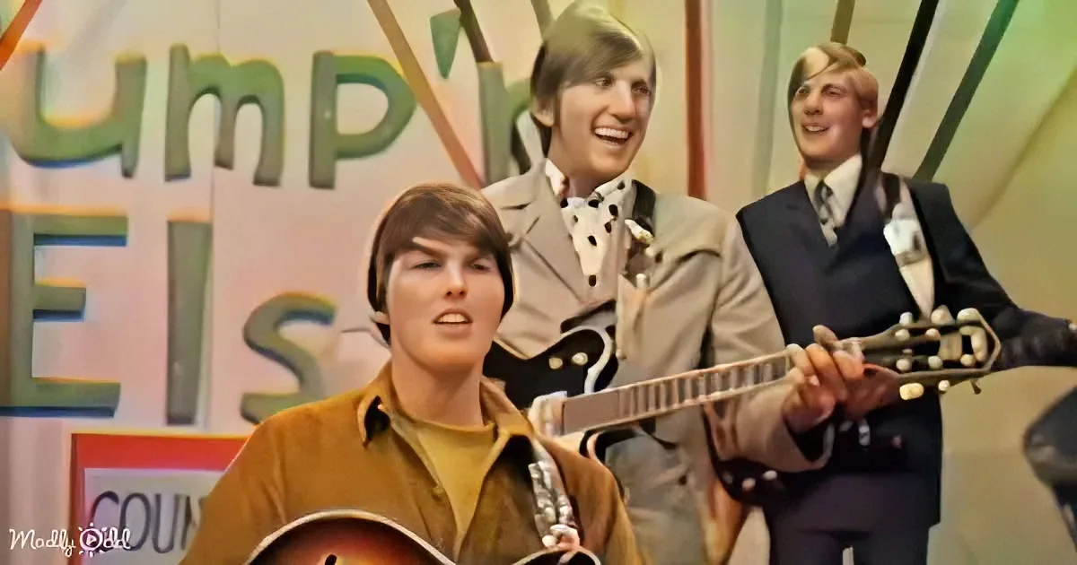 A man in a brown shirt holds a guitar, singing, with two others in suits behind him, smiling.