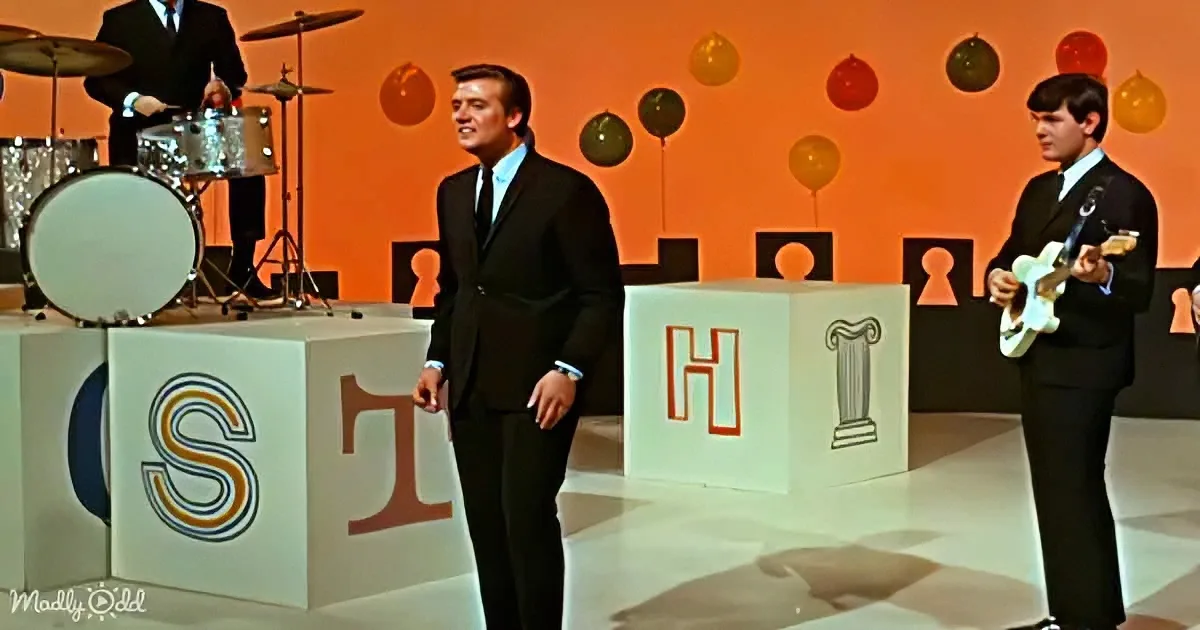A man in a black suit stands on a stage with large blocks spelling "S-T-H," while a guitarist and drummer perform behind him.