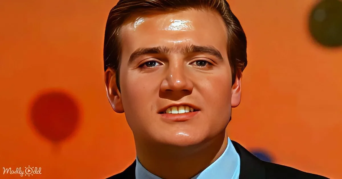 A man in a suit with a bright orange background and a "Madly Odd" watermark.