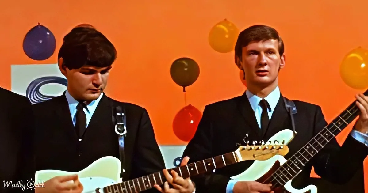 Two men in suits play guitars against an orange backdrop with balloons.