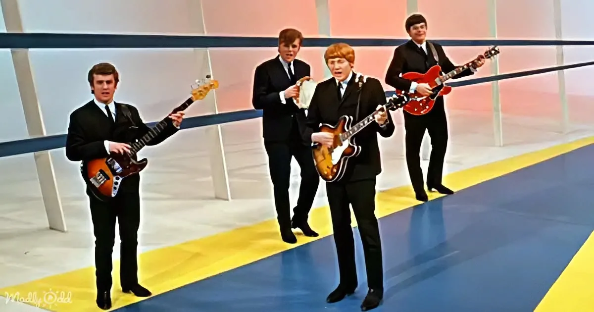 Four musicians in suits play guitars on a stage with a blue and yellow backdrop.