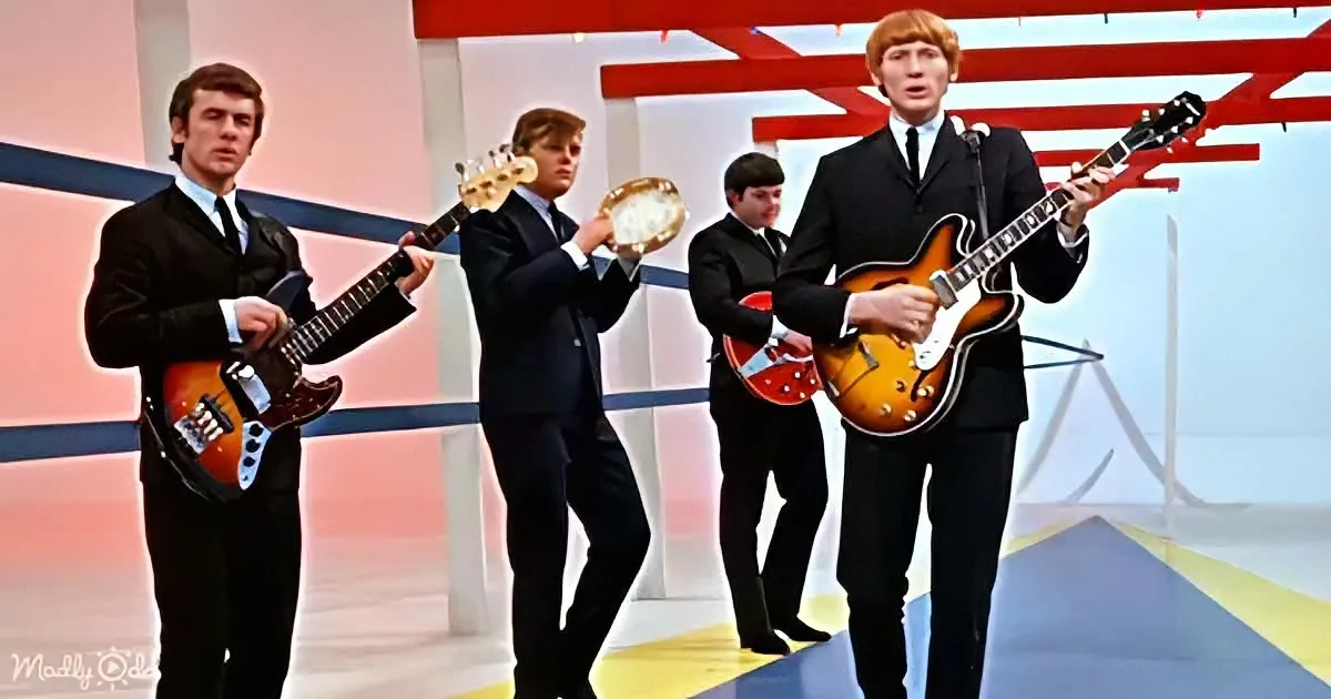 A man in a black suit plays a guitar on stage with other musicians.