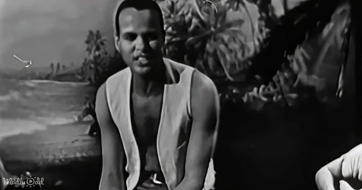 A man in a white vest sits outdoors, with tropical foliage and a body of water in the background.