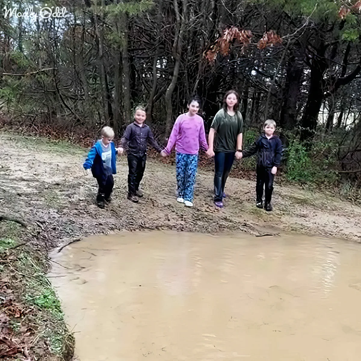 Five children hold hands, standing near a muddy puddle in a forested area.