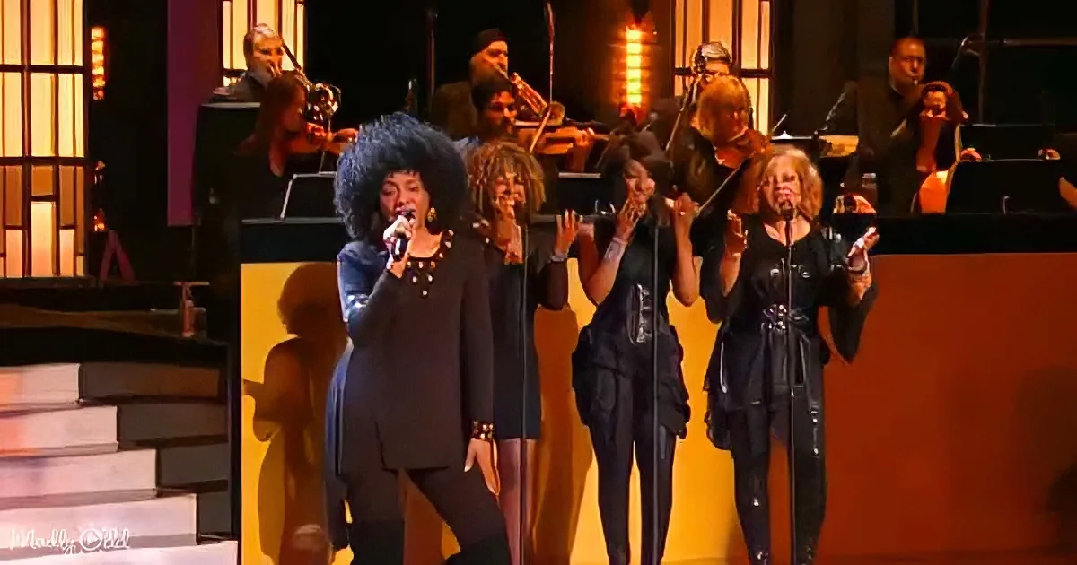 A woman in a black outfit sings into a microphone on stage with other performers and an orchestra behind her.
