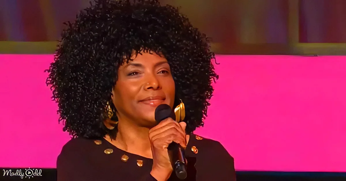 A woman with curly hair holds a microphone, wearing a black outfit with gold buttons, against a pink background.