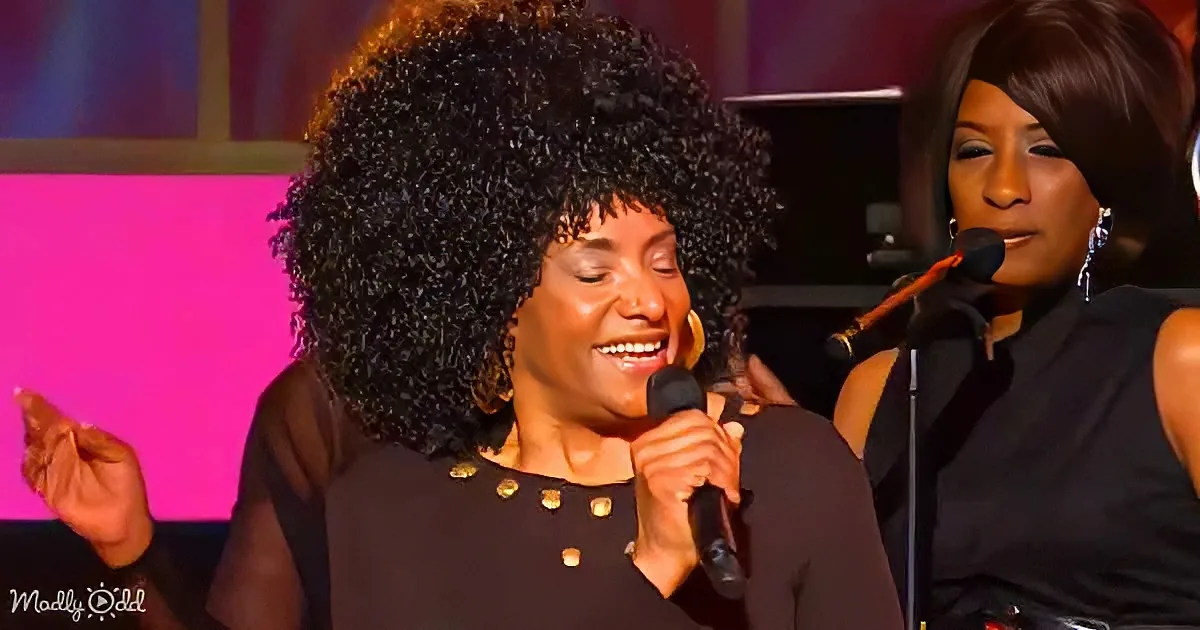 A woman with a large afro hairstyle sings into a microphone on stage, accompanied by another performer.