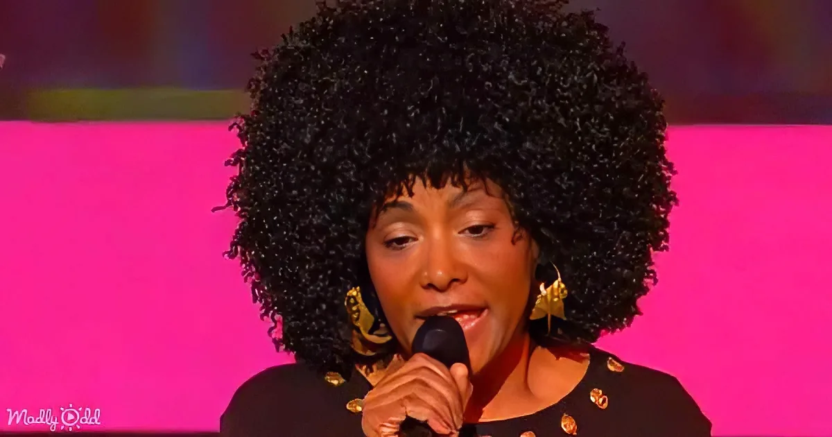 A woman with a large, curly afro hairstyle is singing into a microphone, wearing a black outfit with gold embellishments.