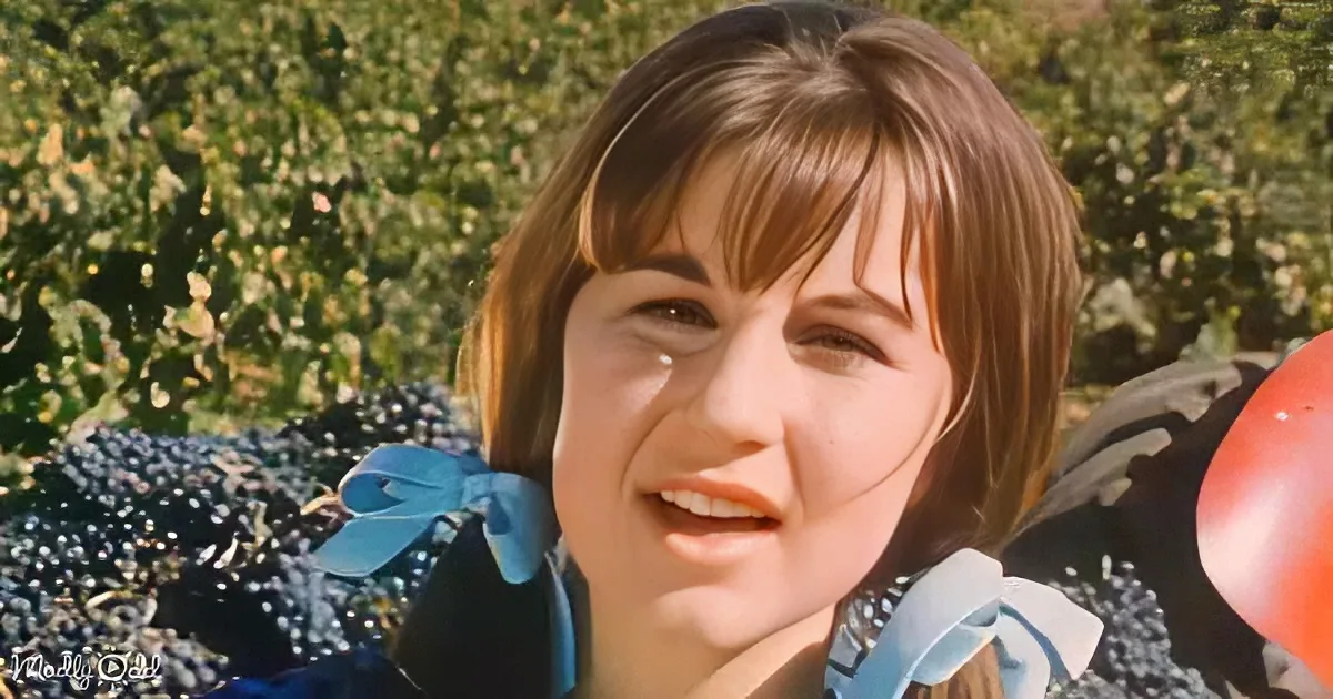 A smiling woman with short brown hair, wearing a blue and white outfit, stands outdoors with greenery in the background.