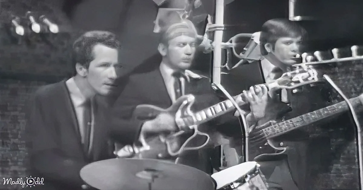 A guitarist in a suit plays an electric guitar, surrounded by other musicians in a band setting.