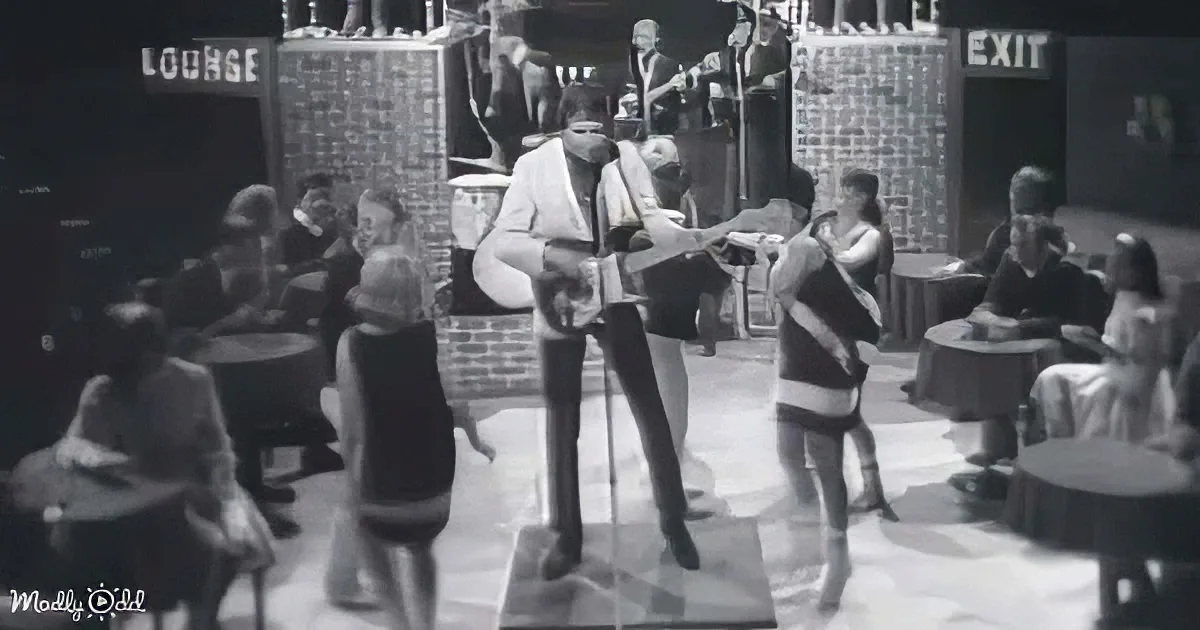 A man in a suit and tie dances on a stage in a dimly lit room with tables and chairs.