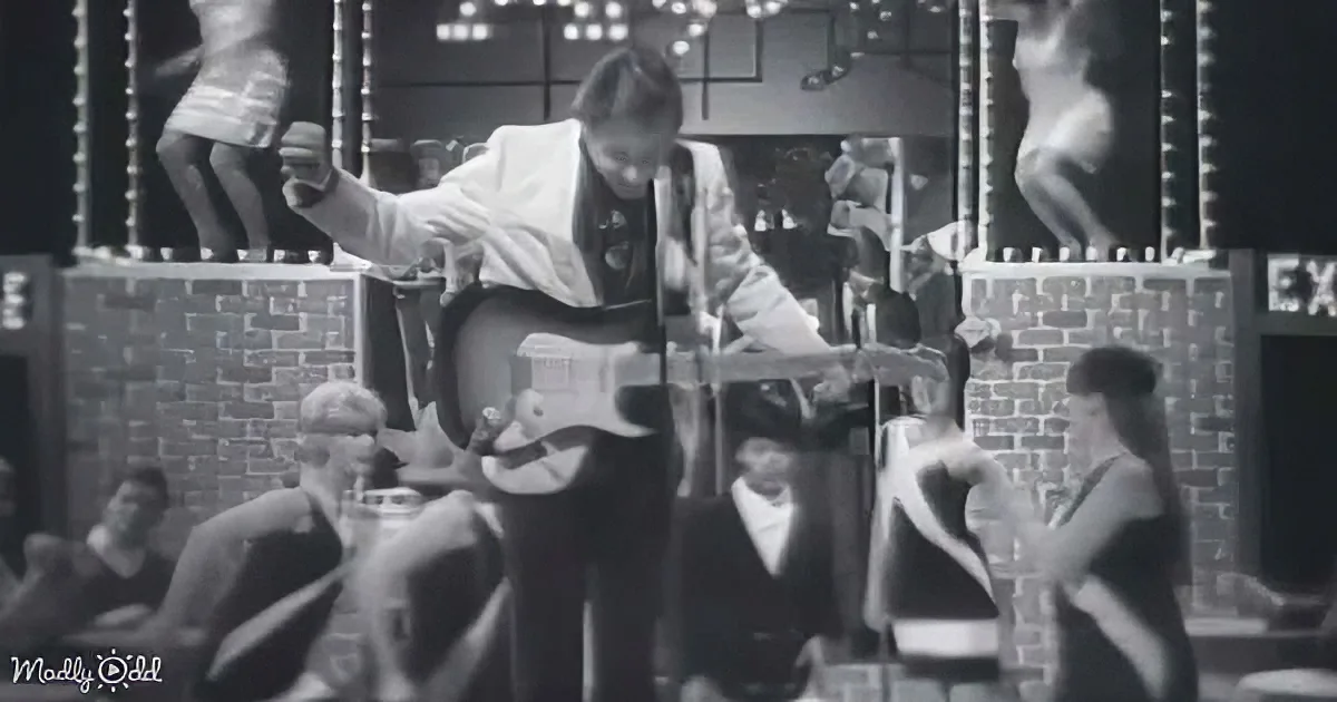 A man in a white suit plays guitar on stage with dancers in the background.
