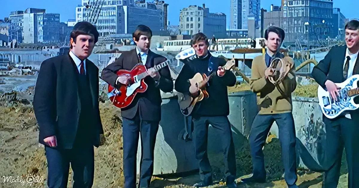 A man in a suit stands in front of a group of musicians playing guitars on a beach.