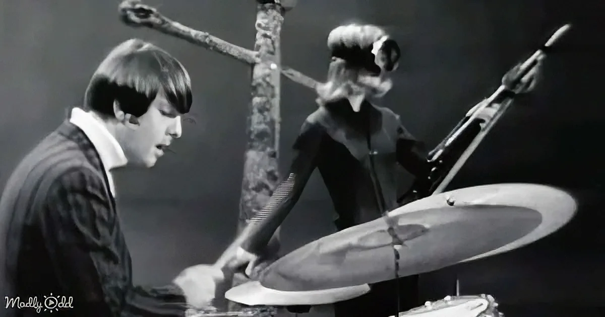 A man in a suit plays piano while a woman in a black dress plays drums, both focused on their instruments.
