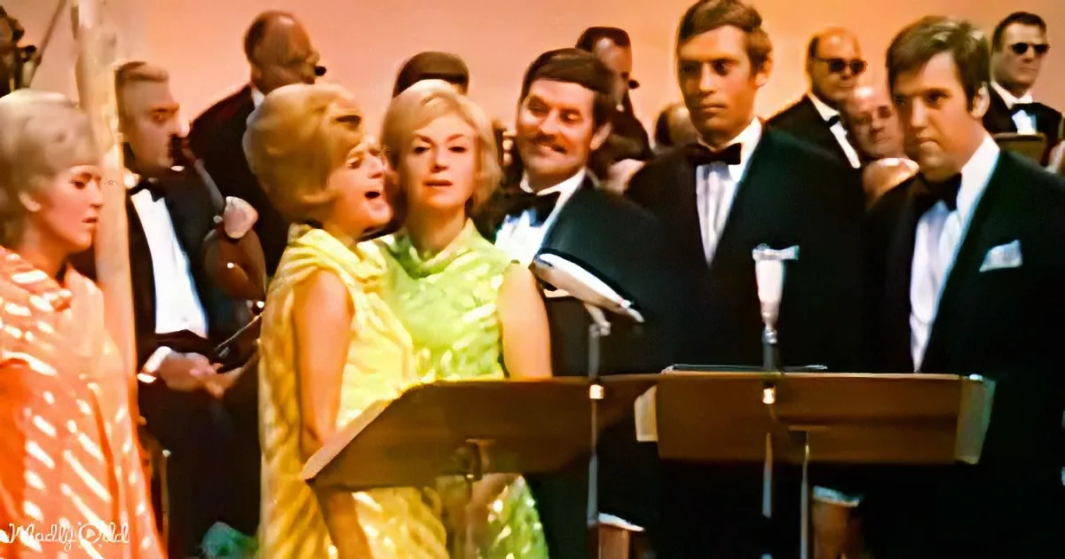 A woman in a yellow dress sings at a podium with a microphone, surrounded by a formal audience in tuxedos.