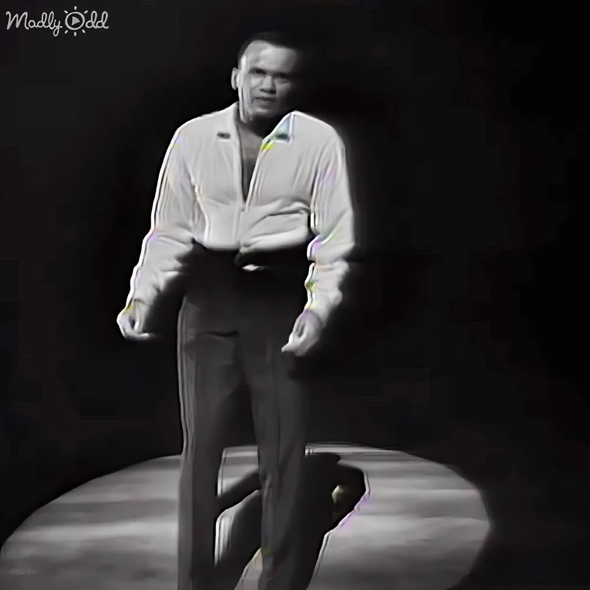 A man in a white shirt and dark pants stands on a platform, illuminated against a dark background.