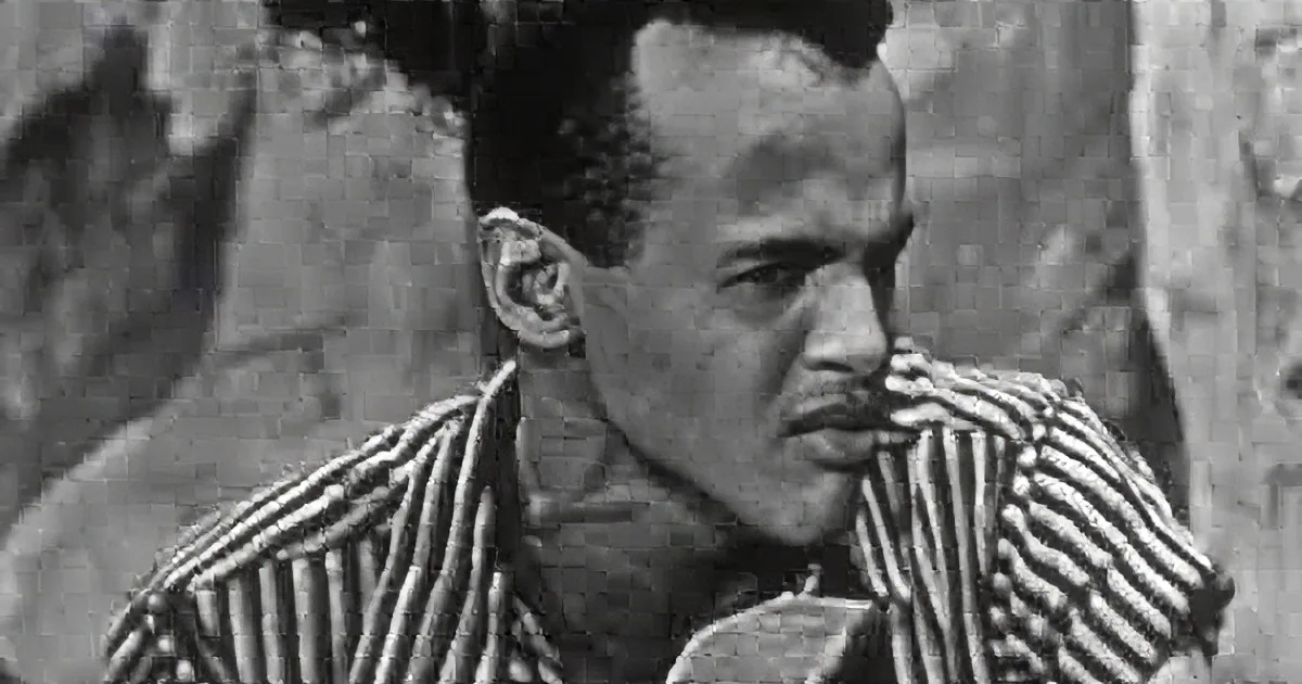 A man in a striped garment is speaking, with a rocky background and a dark, possibly smoky, cloud above.