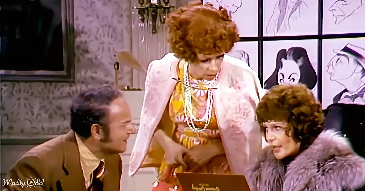 A woman in a colorful dress and pearls stands between two others, one in a fur coat, in a room with sketches on the wall.