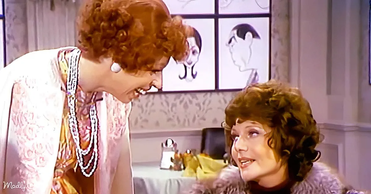 A woman in a floral dress leans over another woman with curly hair, both in a room with a window showing a dog and a person.