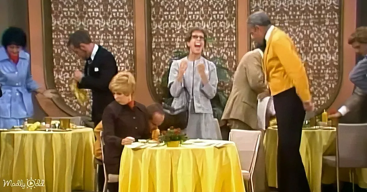 A man in a light gray suit is laughing heartily, surrounded by people in a restaurant setting with yellow tablecloths.