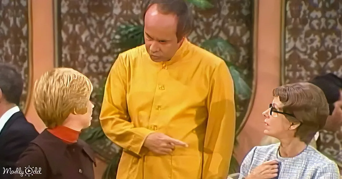 A man in a yellow robe points while standing between two seated individuals in a room with patterned wallpaper.