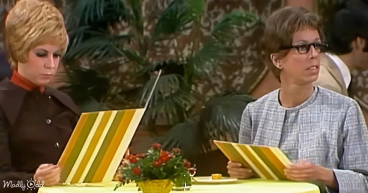 Two women sit at a table, each holding a striped menu, with a floral centerpiece and patterned wallpaper in the background.