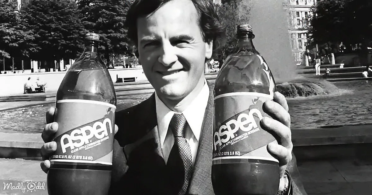 A man in a suit holds two bottles of Aspen soda, smiling in a park setting.