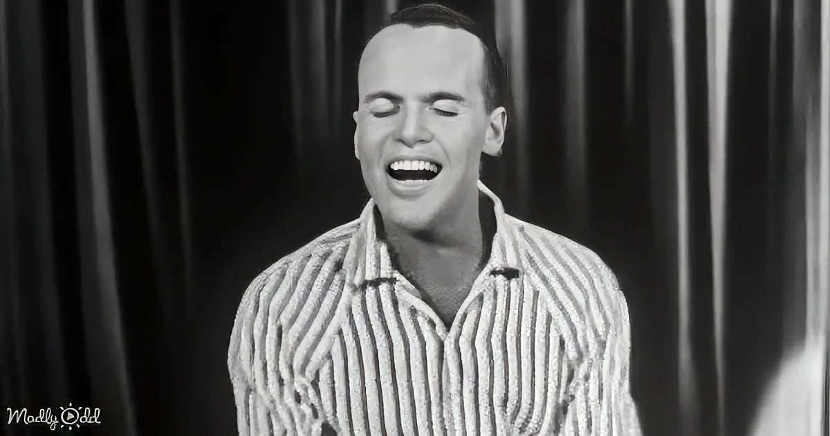 A man in a striped shirt is laughing with his eyes closed, standing in front of a dark curtain.