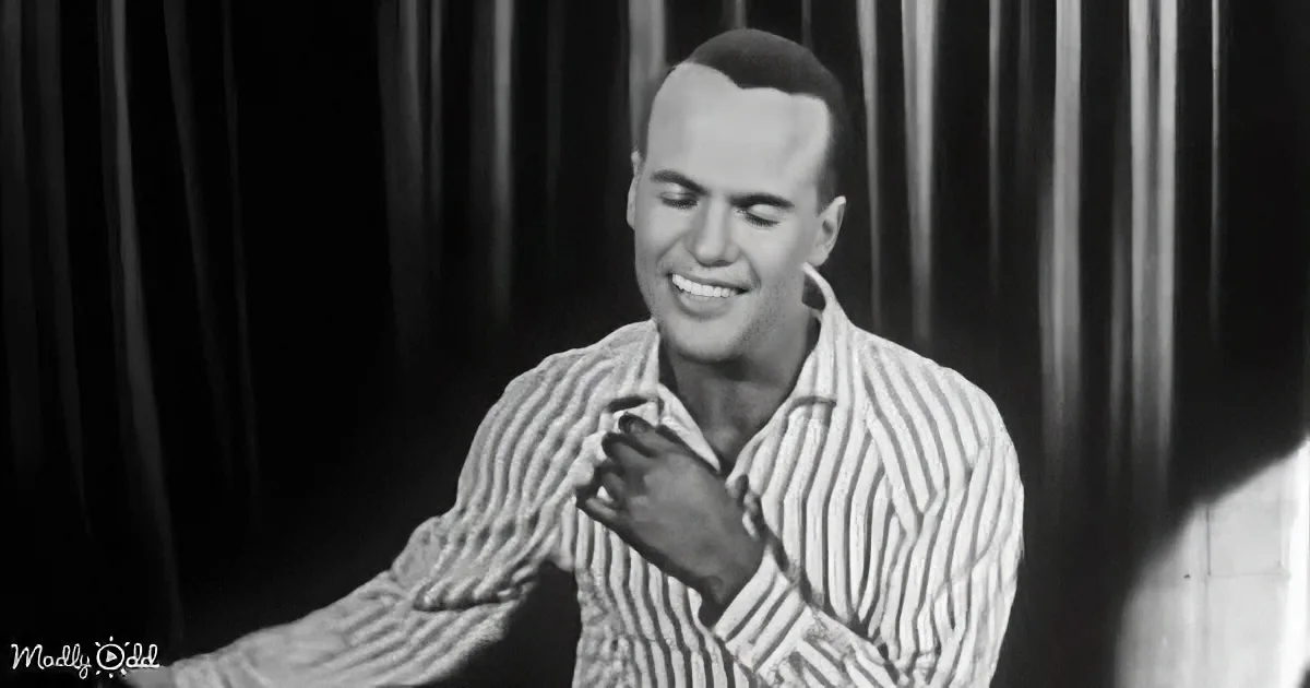 A man in a striped shirt smiles, hands clasped near his chest, against a dark curtain backdrop.