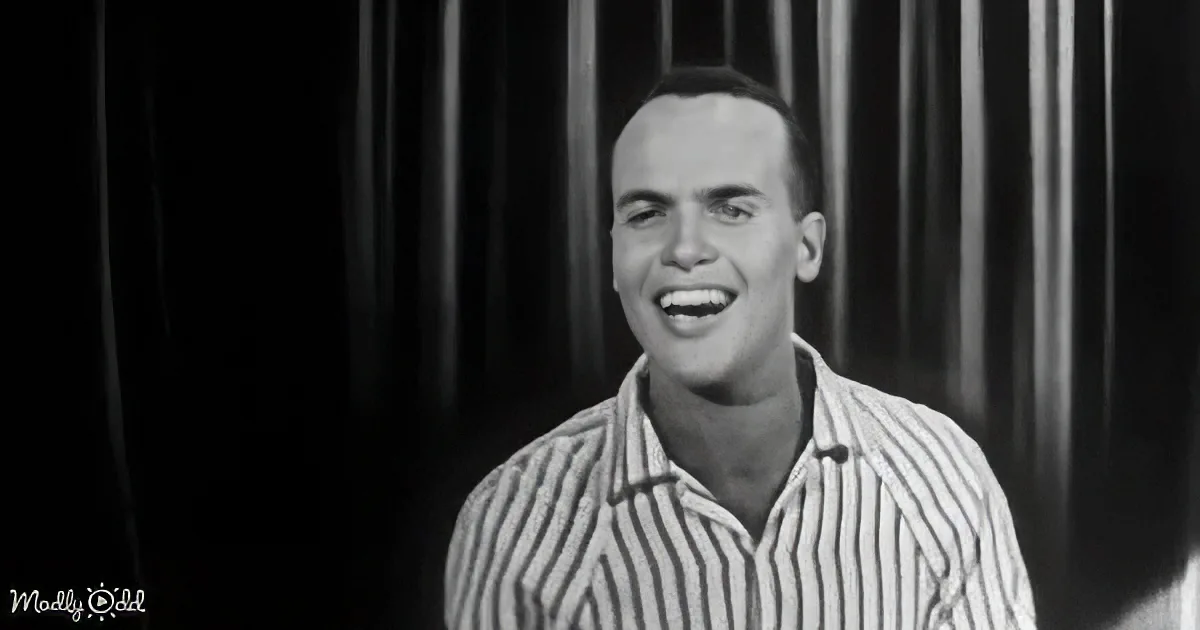 A man in a striped shirt smiles widely, standing in front of dark vertical blinds.