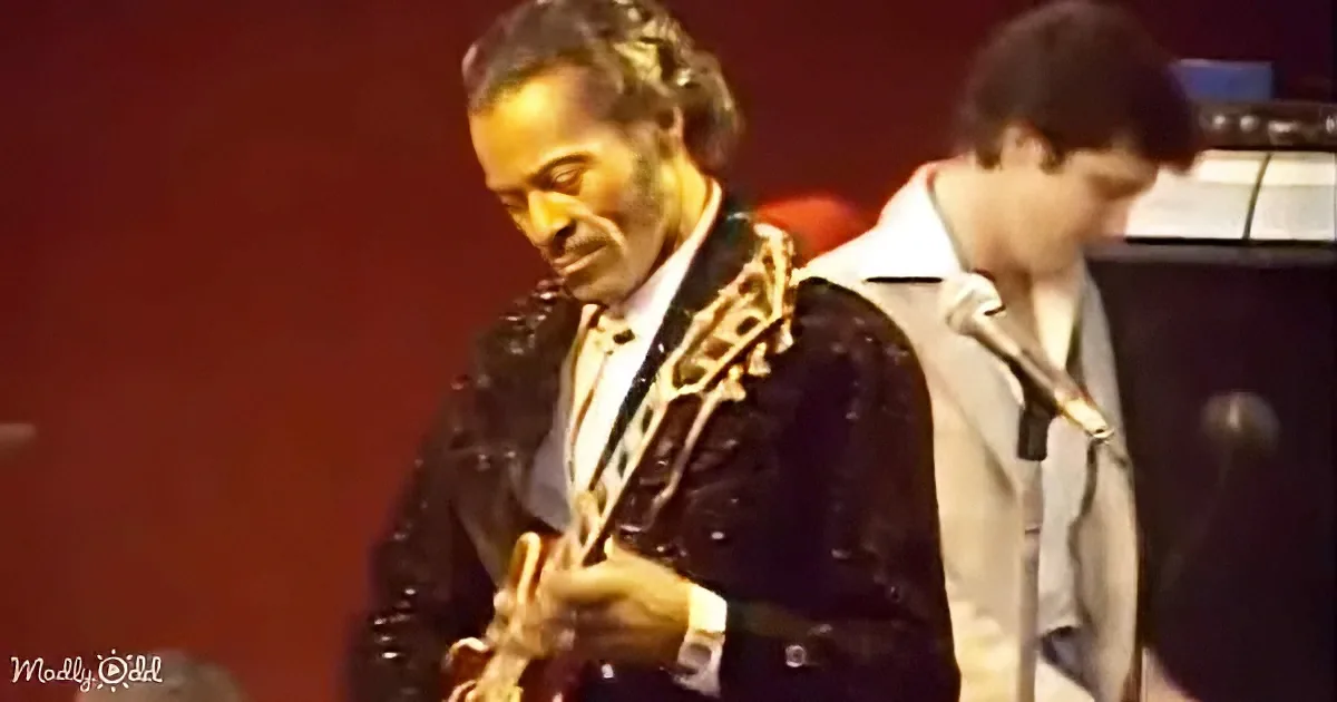 A man in a black jacket plays a guitar on stage, with another person in the background.